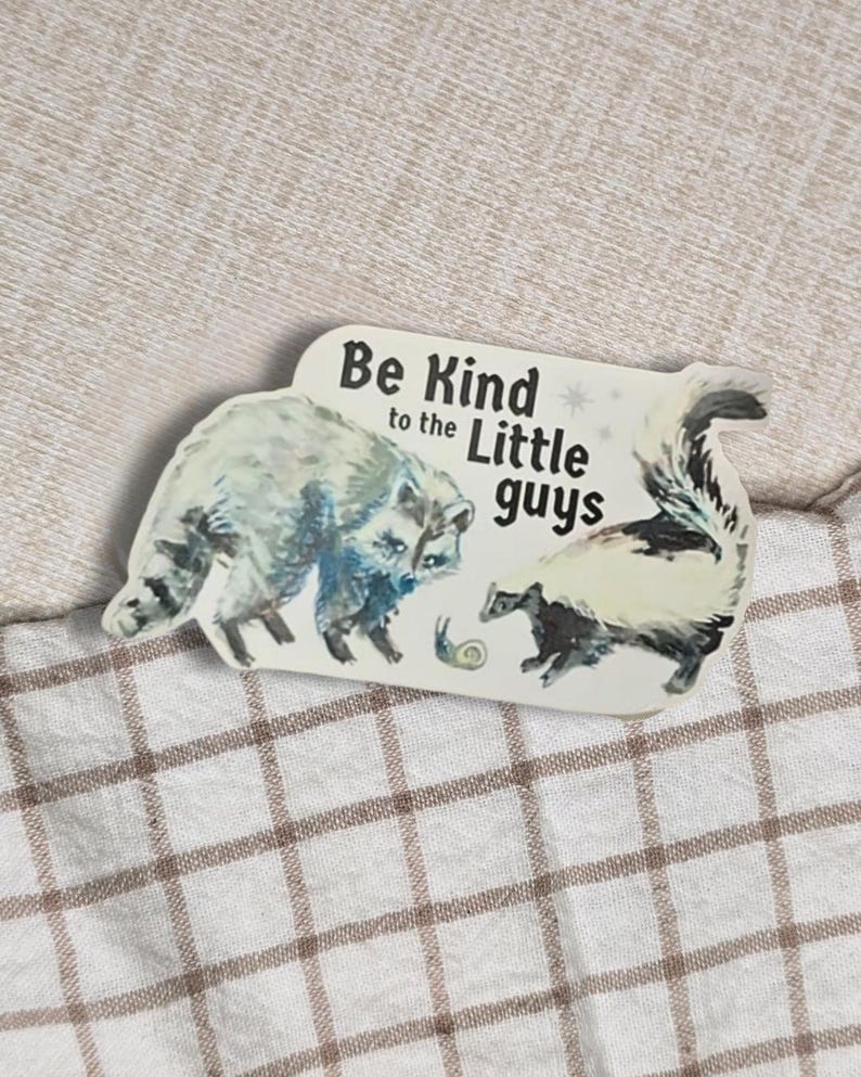 May include: A rectangular sticker featuring a raccoon and a skunk, with the text "Be Kind to the Little Guys." The raccoon is depicted in shades of blue and gray, while the skunk is black and white. A small snail is also present.