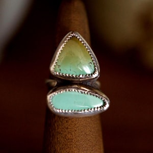Puede incluir: Anillo de plata con dos piedras turquesas. La piedra superior es triangular, la inferior en forma de l&aacute;grima. Las piedras son de color verde claro y est&aacute;n engastadas en plata. Se muestra en un soporte de madera.