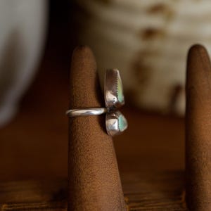 Puede incluir: Un anillo de plata con dos piedras de color turquesa. El anillo se muestra en un soporte de madera. Las piedras tienen un borde plateado y una superficie texturizada. El anillo es una pieza de joyer&iacute;a &uacute;nica.