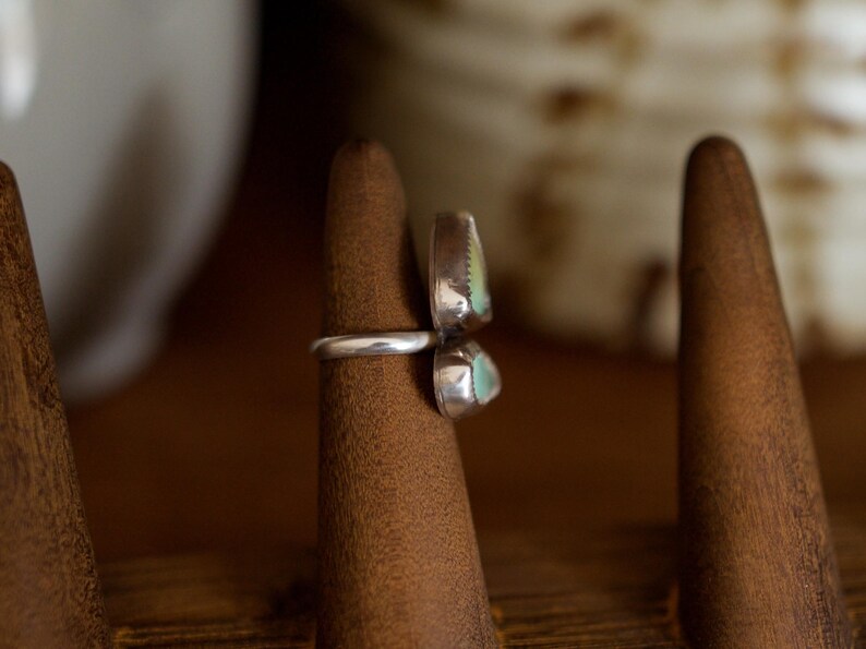 Puede incluir: Un anillo de plata con dos piedras de color turquesa. El anillo se muestra en un soporte de madera. Las piedras tienen un borde plateado y una superficie texturizada. El anillo es una pieza de joyer&iacute;a &uacute;nica.