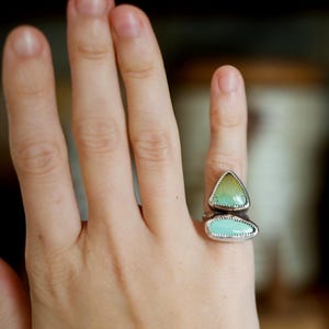 Puede incluir: Un anillo de plata con dos piedras de color turquesa. La piedra superior tiene forma de tri&aacute;ngulo con un tono amarillo-verdoso, y la piedra inferior es un rect&aacute;ngulo redondeado. El anillo est&aacute; en una mano.