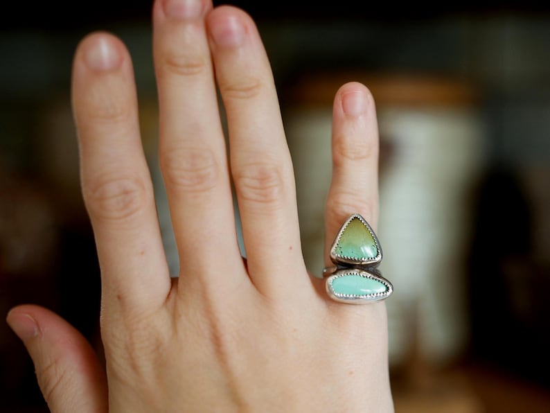 Puede incluir: Un anillo de plata con dos piedras de color turquesa. La piedra superior tiene forma de tri&aacute;ngulo con un tono amarillo-verdoso, y la piedra inferior es un rect&aacute;ngulo redondeado. El anillo est&aacute; en una mano.