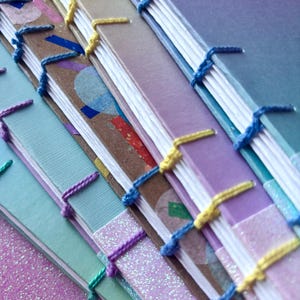 May include: A collection of handmade journals with vibrant covers and hand-stitched spines. The covers are in shades of blue, purple, and brown, with decorative elements and glitter. The binding thread is in matching colours.
