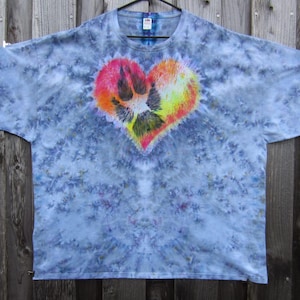 May include: A tie-dye t-shirt with a heart-shaped design. The heart features a paw print in the center, with a gradient of colors including pink, orange, yellow, and black. The shirt has a blue and white tie-dye pattern.