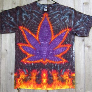 May include: Tie-dye t-shirt with a large purple cannabis leaf design outlined in orange. The shirt has a dark background with blue and black tie-dye patterns. The bottom of the shirt features an orange and yellow flame design.