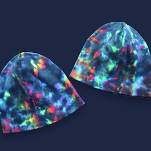 May include: Two tie-dye beanies in a dark blue background. The beanies have a vibrant pattern of neon green, pink, orange, and yellow colors. The beanies are made of a soft material and are suitable for cold weather.