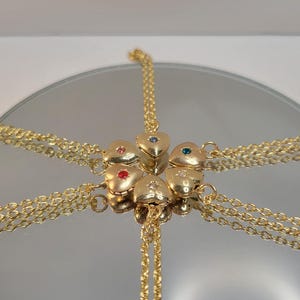 May include: Gold-toned heart-shaped pendants with gemstone accents, connected by delicate chains. The pendants are arranged around a central point, reflecting on a mirrored surface. The gemstones are red, blue, and green.