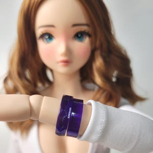 May include: Two purple resin rings are displayed on a doll's arm. One ring has a faceted design, while the other is smooth. The rings are a deep, translucent purple, and the doll is wearing a white top.