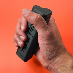 Puede incluir: Un agarre negro con el texto "YOOM TEX" se sostiene en una mano sobre un fondo naranja. El agarre tiene una parte superior en forma de corazón y una superficie texturizada.