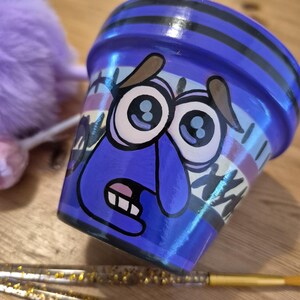 Puede incluir: Una maceta de cerámica azul pintada a mano con un diseño de cara de dibujos animados. La maceta presenta una expresión de sorpresa con ojos grandes, boca muy abierta y cejas marrones. Rayas negras y detalles de estilo graffiti decoran la maceta.