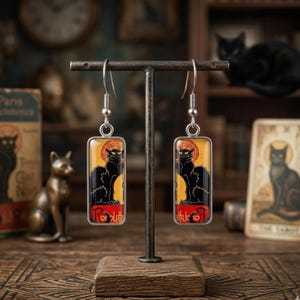 May include: A pair of silver-toned dangle earrings featuring a black cat design. Each earring has a rectangular pendant with a black cat silhouette against a yellow and red background. The earrings are displayed on a brown stand.