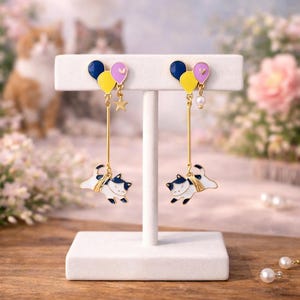 May include: A pair of dangle earrings featuring a cat design. The earrings have a gold-colored stem with a white cat with black markings at the bottom. Above the cat are three colorful balloons in yellow, purple, and blue. A star and a pearl are also included.