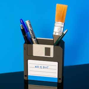 Könnte beinhalten: Ein einzigartiger Schreibtisch-Organizer aus einer wiederverwendeten Diskette. Er enthält Stifte, einen Pinsel und einen Bleistift. Das Etikett lautet "WIN 95 BOOT". Der Hintergrund ist leuchtend blau.