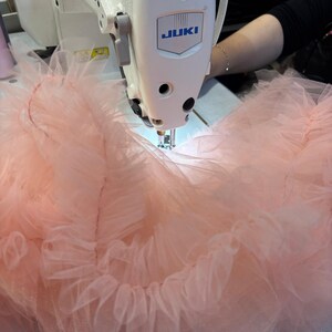 Puede incluir: Primer plano de una m&aacute;quina de coser Juki blanca cosiendo tela rosa claro con volantes. La tela parece ser tul o un material transparente similar. El logotipo de la m&aacute;quina es azul. La imagen muestra la costura de una prenda.