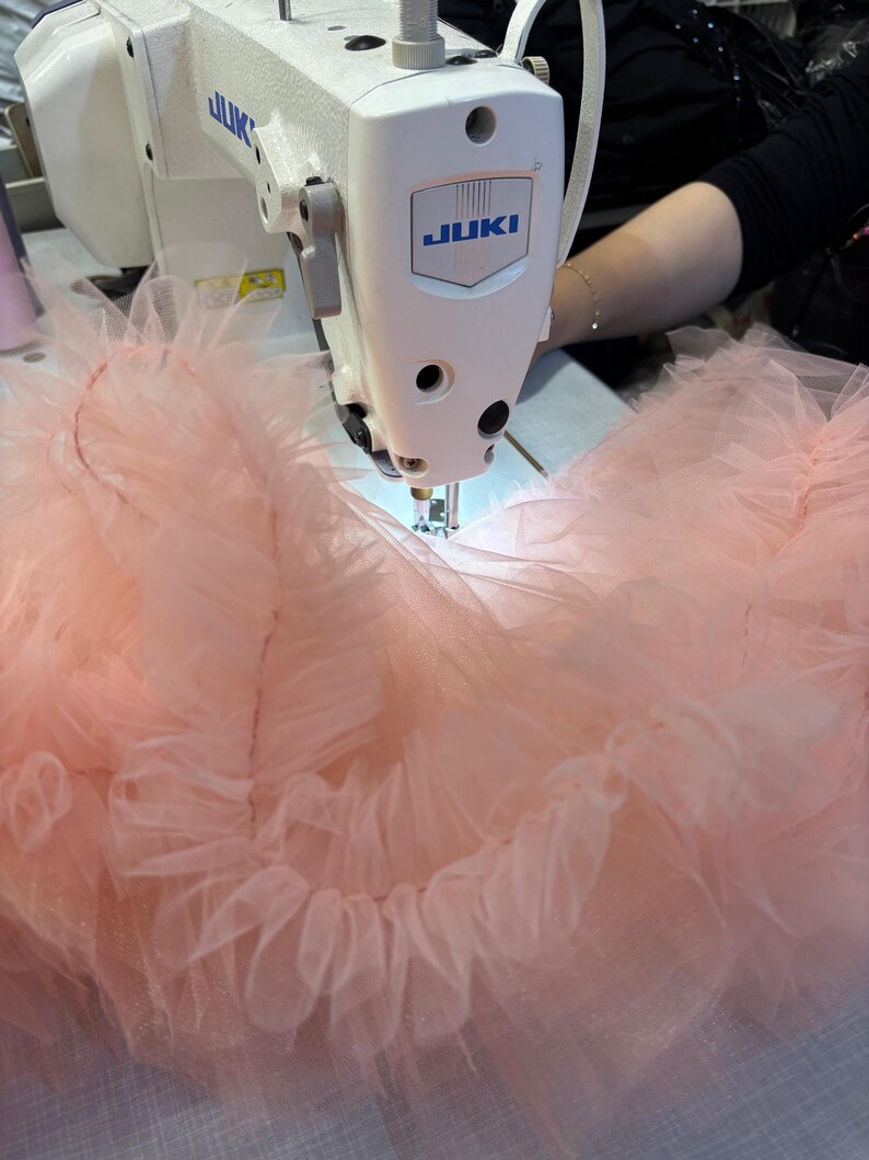 Puede incluir: Primer plano de una m&aacute;quina de coser Juki blanca cosiendo tela rosa claro con volantes. La tela parece ser tul o un material transparente similar. El logotipo de la m&aacute;quina es azul. La imagen muestra la costura de una prenda.