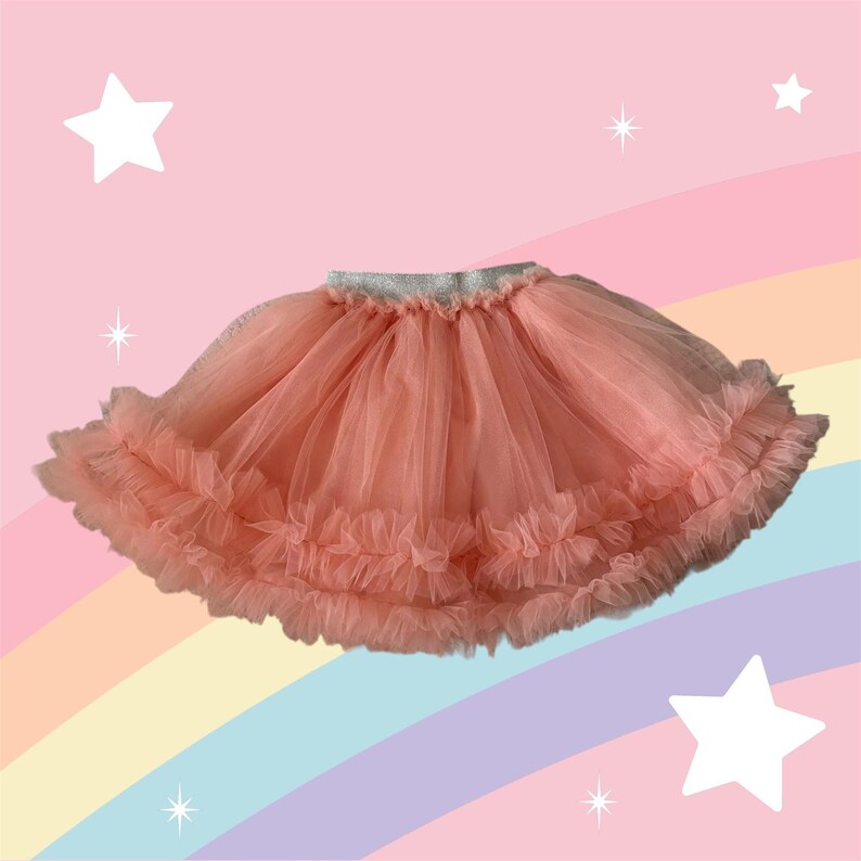 Puede incluir: Una falda tut&uacute; de color coral con m&uacute;ltiples capas de tul y un dobladillo con volantes. La cintura es plateada. El fondo es un arco&iacute;ris pastel con estrellas blancas.