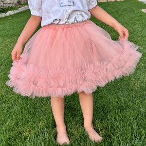 Puede incluir: Una falda de tul rosa claro con un dobladillo con volantes. La falda la lleva una persona de pie sobre hierba verde. La persona lleva una camiseta blanca con un dise&ntilde;o.