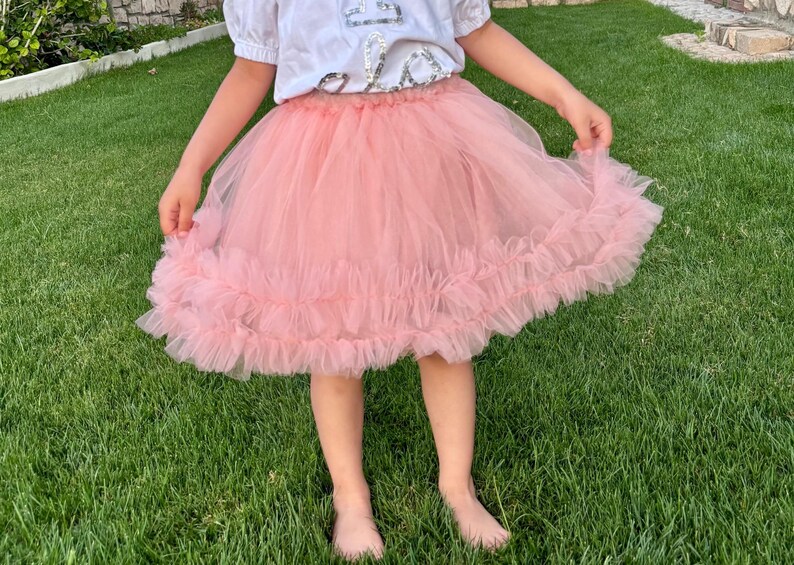 Puede incluir: Una falda de tul rosa claro con un dobladillo con volantes. La falda la lleva una persona de pie sobre hierba verde. La persona lleva una camiseta blanca con un dise&ntilde;o.