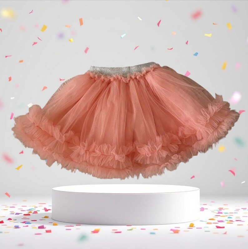 Puede incluir: Una falda tut&uacute; de color coral con m&uacute;ltiples capas de tul suave. La falda tiene una cintura plateada y bordes con volantes. La falda se exhibe sobre un pedestal blanco con confeti colorido en el fondo.