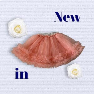 Puede incluir: Falda de tul color coral con dobladillo con volantes, sobre un fondo blanco texturizado. Las palabras "New" e "in" se muestran encima y debajo de la falda. Dos rosas blancas se colocan a cada lado.