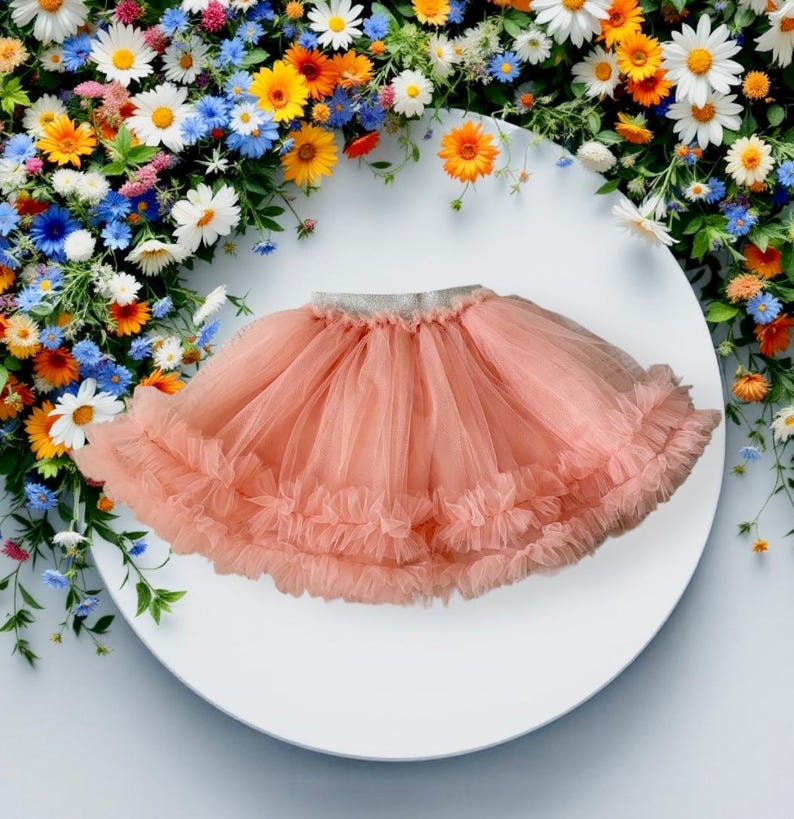 Puede incluir: Una falda de tul color melocot&oacute;n con m&uacute;ltiples capas y un dobladillo con volantes. La falda tiene una cintura plateada y se muestra sobre una superficie blanca, rodeada de un vibrante arreglo floral de flores coloridas, incluidas margaritas.