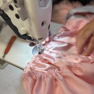 Puede incluir: Primer plano de una m&aacute;quina de coser Juki en acci&oacute;n, cosiendo tela de sat&eacute;n rosa claro. La tela est&aacute; fruncida, creando un efecto de volantes. La m&aacute;quina de coser es blanca y tiene el logotipo de Juki. Escena de costura.
