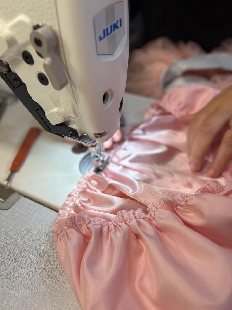 Puede incluir: Primer plano de una m&aacute;quina de coser Juki en acci&oacute;n, cosiendo tela de sat&eacute;n rosa claro. La tela est&aacute; fruncida, creando un efecto de volantes. La m&aacute;quina de coser es blanca y tiene el logotipo de Juki. Escena de costura.