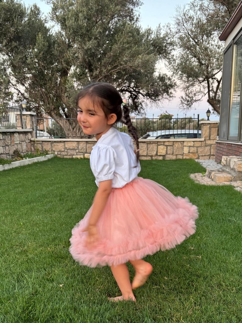 Puede incluir: Una ni&ntilde;a con una camiseta blanca de manga corta y una falda tut&uacute; rosa, girando en un c&eacute;sped verde. La falda es en capas y voluminosa. La ni&ntilde;a tiene el pelo oscuro y est&aacute; descalza.