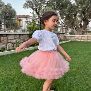 Puede incluir: Una ni&ntilde;a peque&ntilde;a con una camiseta blanca con el n&uacute;mero "1" y una falda tut&uacute; rosa claro. La ni&ntilde;a est&aacute; de pie sobre hierba verde, con &aacute;rboles y un muro de piedra al fondo. La falda es de un material suave y esponjoso.