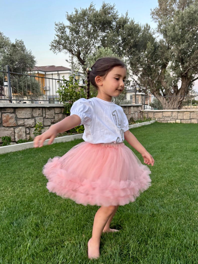 Puede incluir: Una ni&ntilde;a peque&ntilde;a con una camiseta blanca con el n&uacute;mero "1" y una falda tut&uacute; rosa claro. La ni&ntilde;a est&aacute; de pie sobre hierba verde, con &aacute;rboles y un muro de piedra al fondo. La falda es de un material suave y esponjoso.