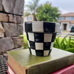 May include: A small, hand-painted flower pot with a black and white checkered pattern. The pot is sitting on top of two vintage books with worn covers. The books are green and red.