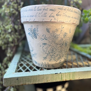 Könnte beinhalten: Cremefarbener Keramik-Blumentopf mit Used-Look. Der Topf hat ein blaues Blumendesign, eine Biene und französischen Schriftzug. Die Worte "LES ROSES" sind auf der Vorderseite aufgedruckt. Der Topf steht auf einem grünen Metallregal.