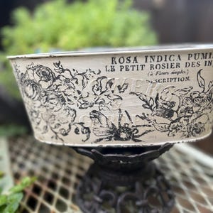 Könnte beinhalten: Cremefarbener Metallpflanzer mit schwarzen Blumen- und Bienenillustrationen und französischem Text. Der oval geformte Pflanzer steht auf einem dekorativen, dunklen Metallständer. Das Design des Pflanzers hat eine Vintage- und Landhausästhetik.