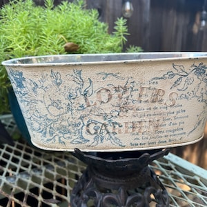 May include: A distressed, rectangular metal planter with blue floral designs and French text, resting on a decorative black stand. The planter is filled with green plants, creating a vintage garden aesthetic.