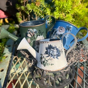 May include: A collection of decorative watering cans in various colors, including white, green, and blue. Each can is adorned with floral patterns. The watering cans are arranged on a metal surface, with greenery in the background.