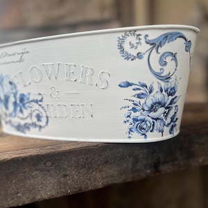 Puede incluir: Jardinera ovalada de metal blanco con diseños florales y de volutas azules. Las palabras "FLOWERS & GARDEN" están grabadas en relieve en la parte delantera. La jardinera está sobre una superficie de madera.