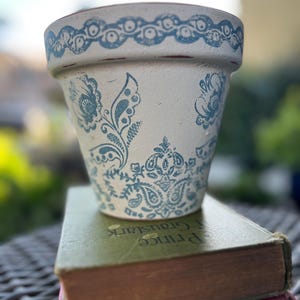 May include: A white ceramic flower pot with a blue floral design. The pot features a decorative blue border around the rim and is sitting on top of two vintage books. The pot is approximately 6 inches tall.