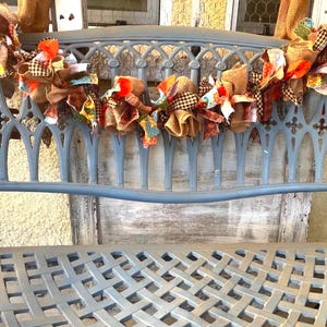 Op de afbeelding: Een decoratieve slinger van stoffen strikken in herfstkleuren, waaronder oranje, bruin en beige, siert de rugleuning van een blauwe metalen bank. De bank heeft een zitting met een roosterpatroon en een sierlijk rugontwerp, wat een rustieke en feestelijke uitstraling creëert.