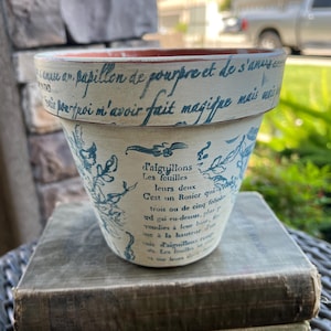 Puede incluir: Una maceta de cerámica color crema con escritura azul y diseños florales. La maceta está apilada encima de tres libros antiguos. La maceta tiene texto en francés alrededor del borde y en el cuerpo.