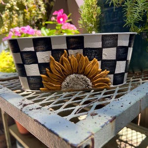 May include: A rectangular planter with a black and white checkered pattern and a 3D sunflower design. The planter is made of ceramic or a similar material and sits on a metal shelf. The background includes greenery and flowers.