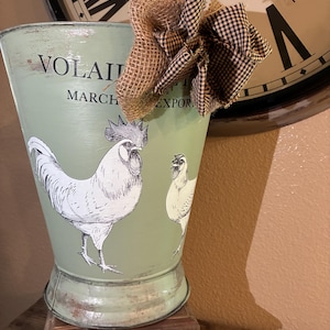 Könnte beinhalten: Ein hellgrüner Metalleimer mit einer Illustration eines Hahns und einer Henne. Auf dem Eimer stehen die Worte "VOLAIL" und "MARCH EXPORT". Eine Schleife aus Jute und schwarz-weißem Karomuster verziert die Oberseite des Eimers.