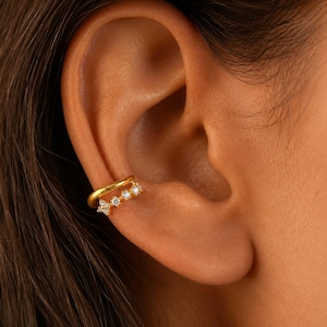 Pode incluir: Um ear cuff de tom dourado com um design de banda dupla. A banda superior é um fio liso e polido de cor dourada, enquanto a banda inferior está cravejada com quatro pequenas pedras claras de corte redondo dispostas em fila na cartilagem.