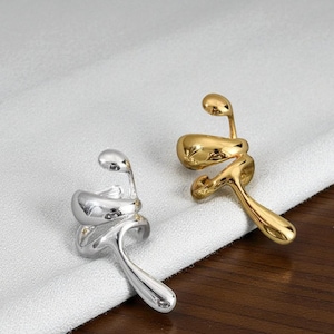 Könnte beinhalten: Zwei abstrakte Ohrringe, einer silberfarben und einer goldfarben. Jeder hat ein fließendes, organisches Design mit einer geschwungenen, wirbelnden Form und einer kleinen, abgerundeten Spitze. Die Ohrringe werden auf einer weißen Stoffoberfläche präsentiert.