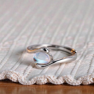 May include: A silver-toned ring with a swirling design, featuring an oval, iridescent gemstone. The ring is set on a white, textured surface, showcasing the jewelry's elegant simplicity. The gemstone reflects a range of colors.