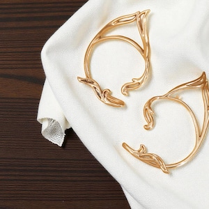 May include: Two gold-coloured ear cuffs with an elven design, featuring curved lines and decorative flourishes. The jewellery is displayed on a white cloth against a dark wood background. The ear cuffs are designed to wrap around the ear.