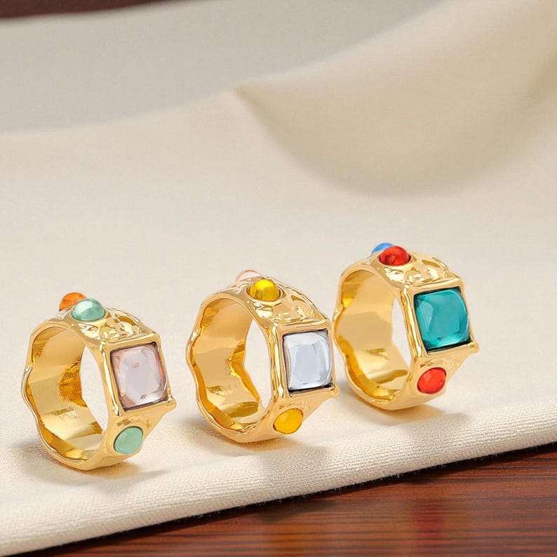 May include: Three gold-colored rings with colorful square and round gemstones. The rings have a textured design and are displayed on a cream-colored surface. The gemstones are in shades of pink, blue, yellow, green, and red.