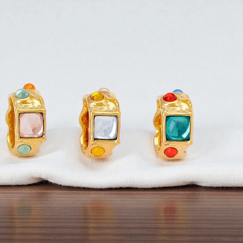 May include: Three gold-toned rings with square, colorful gemstones. Each ring features a different gemstone color: pink, white, and teal. Additional smaller gemstones in orange, yellow, green, and red accent the rings. The rings are displayed on a white cloth.