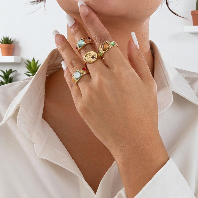 May include: A collection of gold-toned rings with colorful gemstone accents. The rings feature various shapes and designs, including square, round, and textured surfaces. Some rings have multiple small gemstones, while others have a single, larger stone.