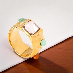 May include: A gold-colored ring with a rectangular, light pink center stone and two smaller, round green stones. An additional orange stone is set at the base. The ring has a textured, organic design.