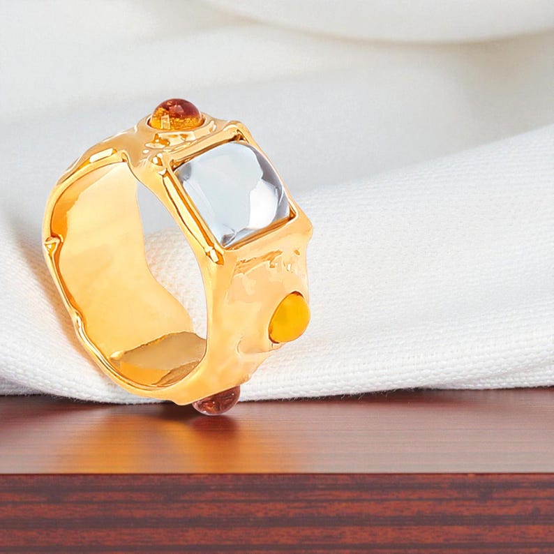 May include: A gold-colored ring with a square, clear gemstone and smaller orange and brown stones. The ring has a thick band and a textured design. The ring is displayed on a white cloth and a wooden surface.